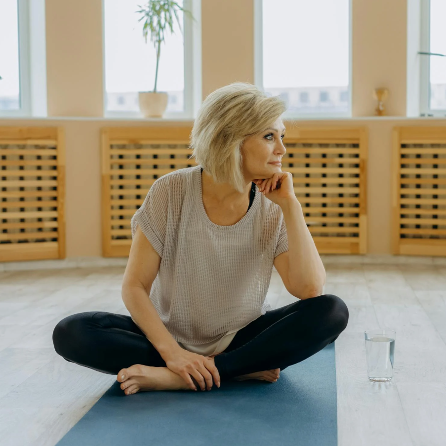 Woman sitting on a yoga mat in a room with large windows and wooden panels.